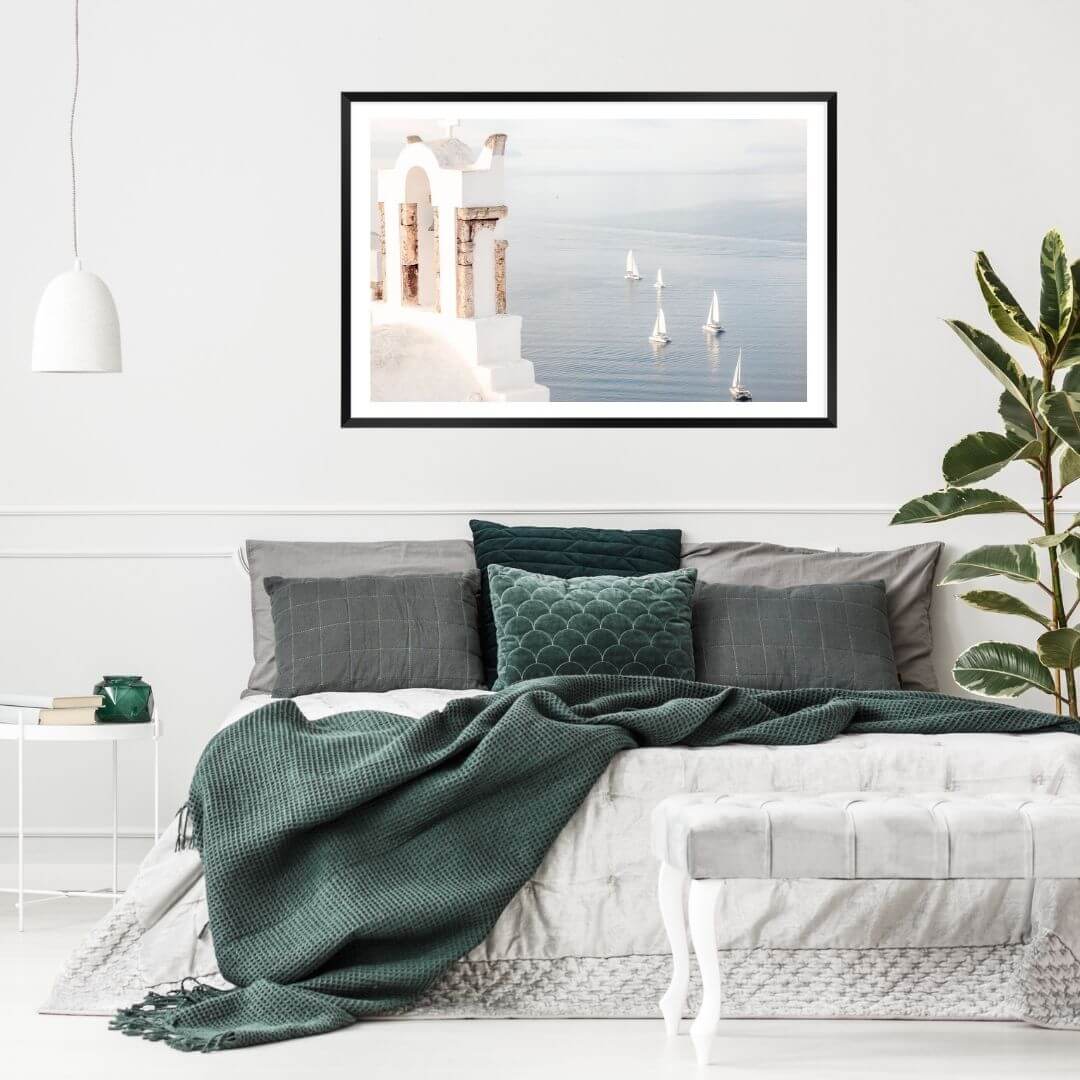 A coastal wall art photo print of Boats sailing on the sea in Santorini Greece  with a black frame, white border on wall above bed