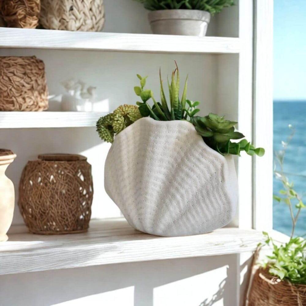 Decorative shelf with a white ceramic oyster vase, woven baskets, and potted plants by a window with a view of water.