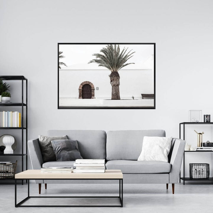 A wall art photo print of a white Spanish Church with a Palm Tree  with a black frame on a wall above a sofa by Beautiful Home Decor