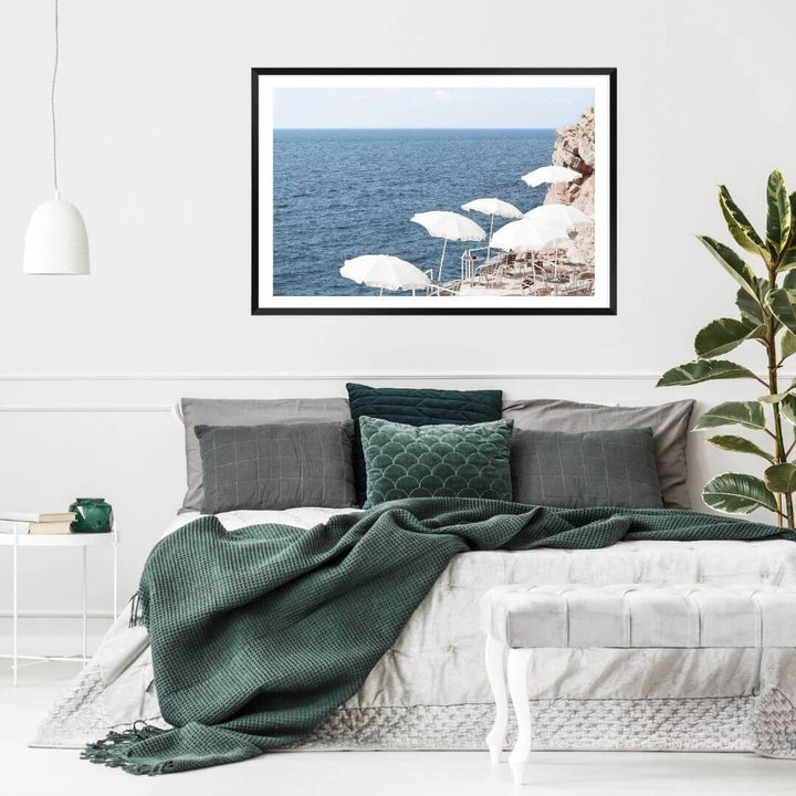 A coastal wall art photo print of white umbrellas on an Amalfi Coast Beach Italy with a black frame, white border on wall above bed