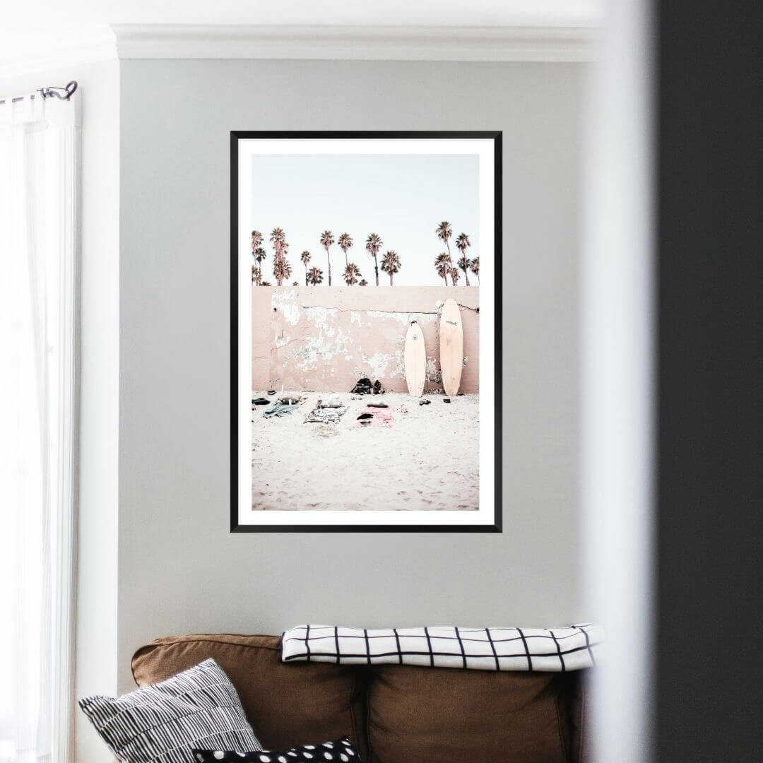 Available framed or unframed, this coastal wall art print features surf boards on the beach with palm trees and a wall.