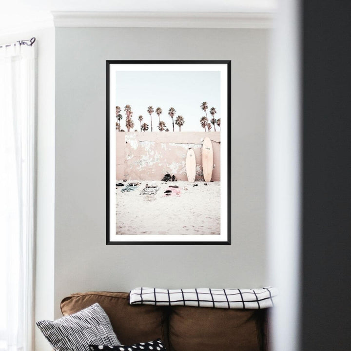 Available framed or unframed, this coastal wall art print features surf boards on the beach with palm trees and a wall.