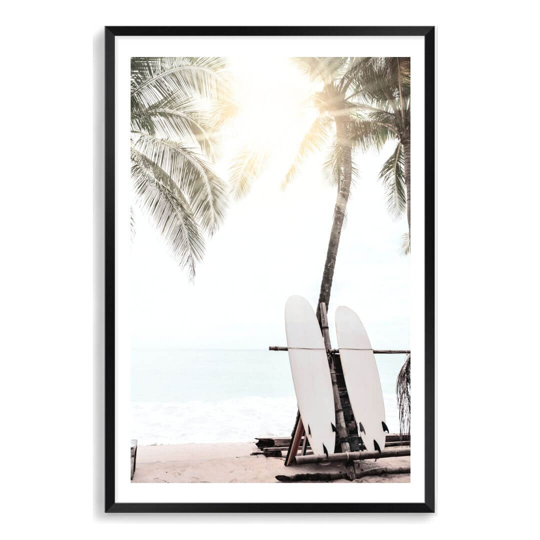 A wall art print of two white surf boards under the palm trees on a surfer beach in Hawaii in a black frame with a white border