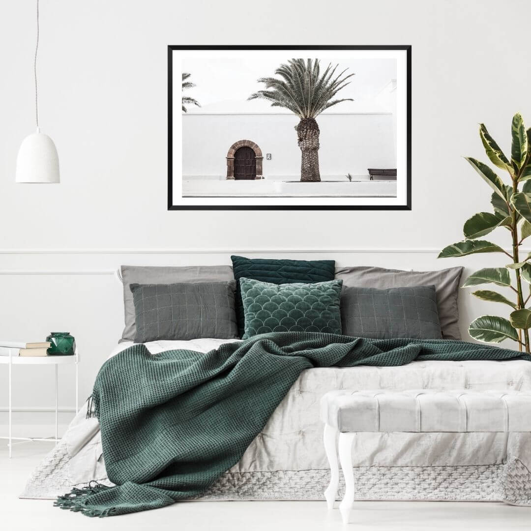 A wall art photo print of a white Spanish Church with a Palm Tree  with a black frame, white border on wall above bed