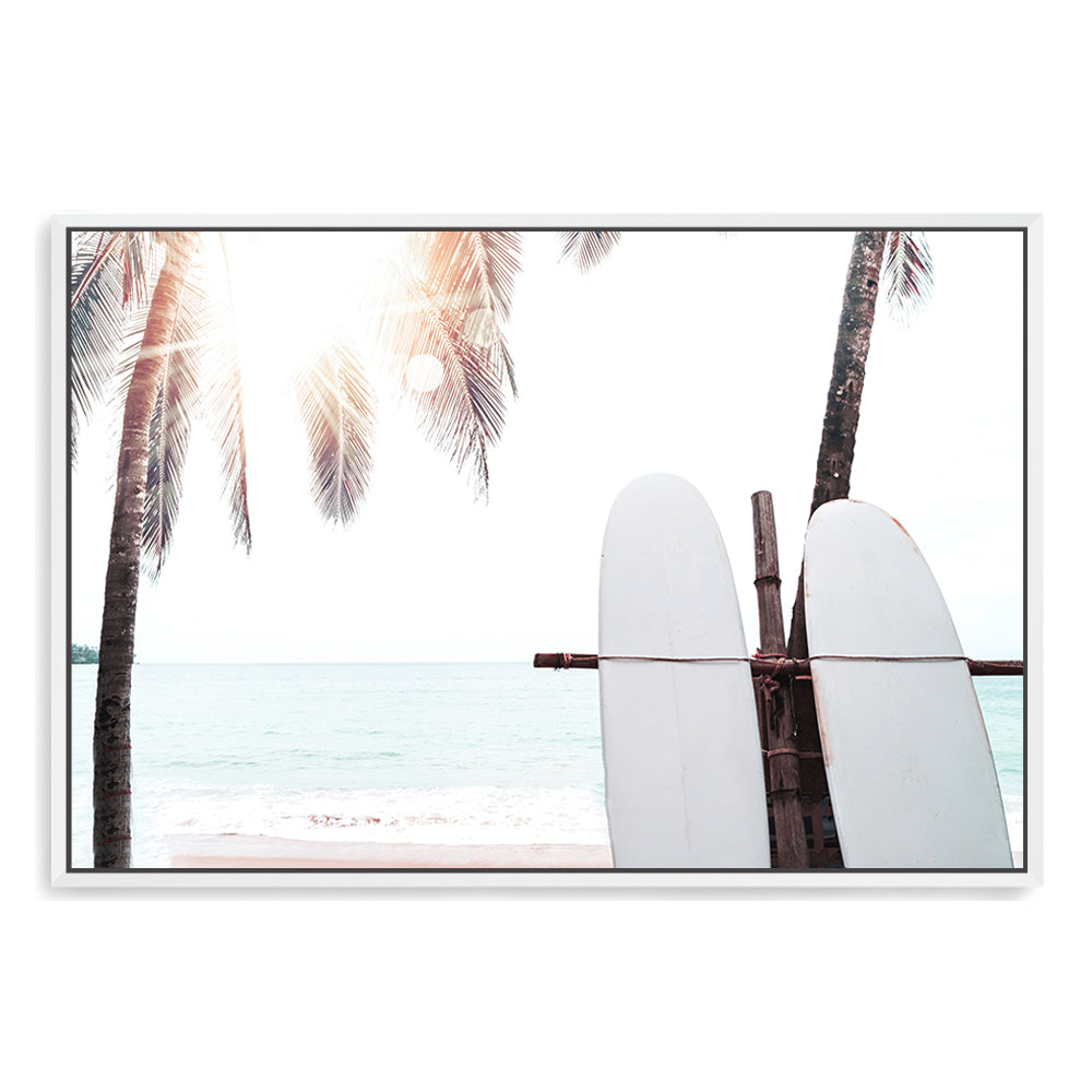 A coastal wall art of two white surfboards on a beach under the palm trees with a tropical sunset view.