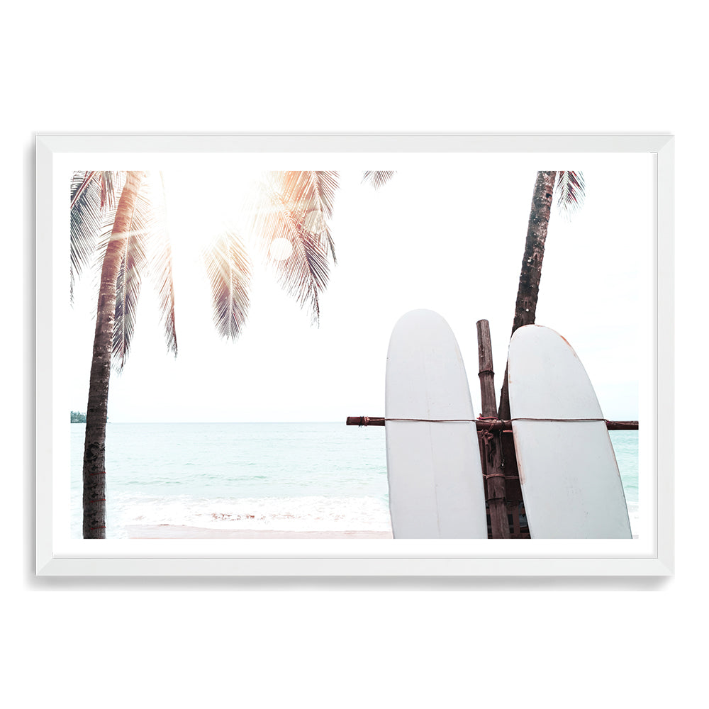 Our Surfers Sunset (A) art prints features two white surf boards on a beach under the palm trees with a view of a tropical sunset.